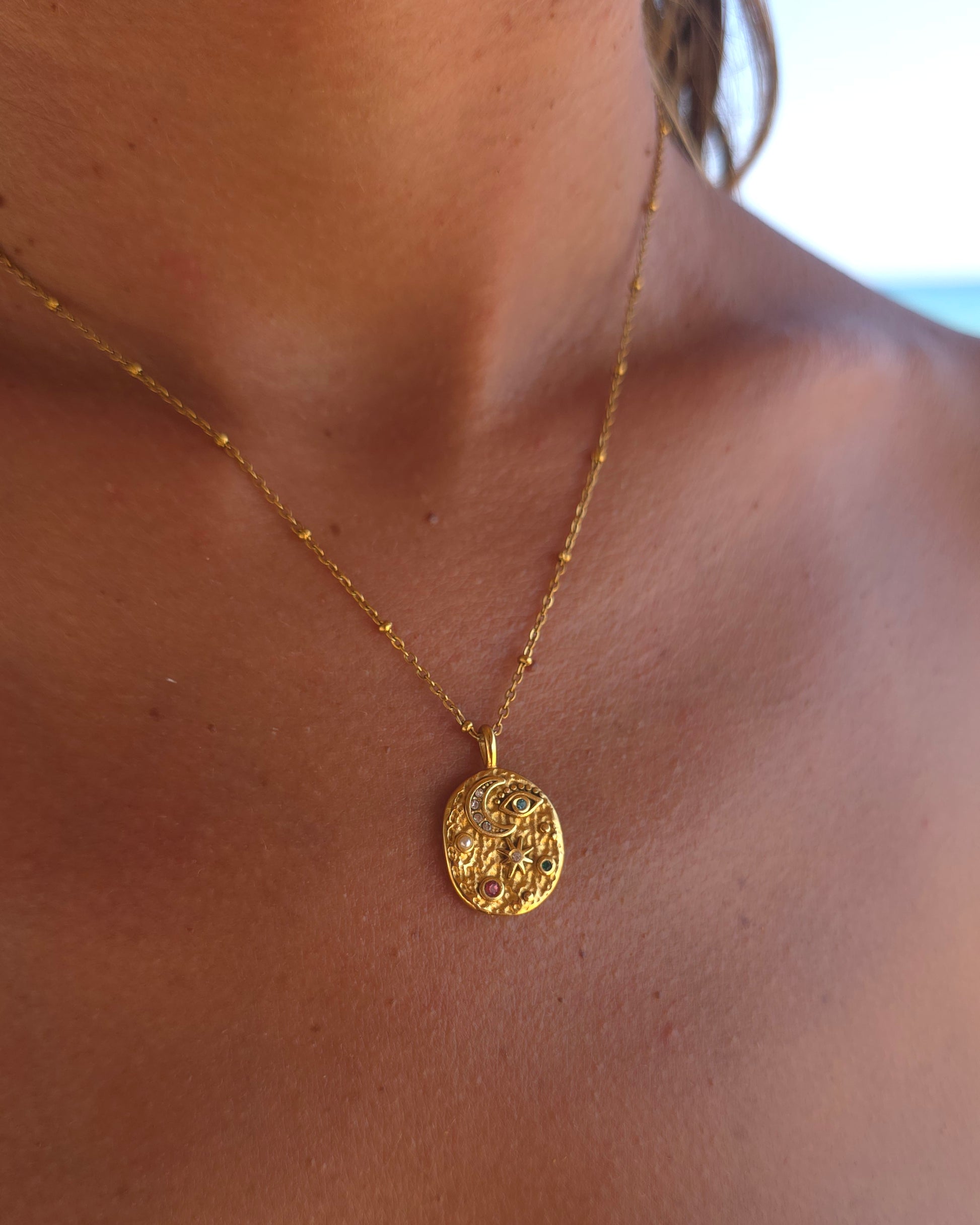 Lifestyle close-up of the gold Celestial necklace in 18k gold plated 316L stainless steel, featuring its symbolic moon and star medallion against sun-kissed skin with a soft beach background.