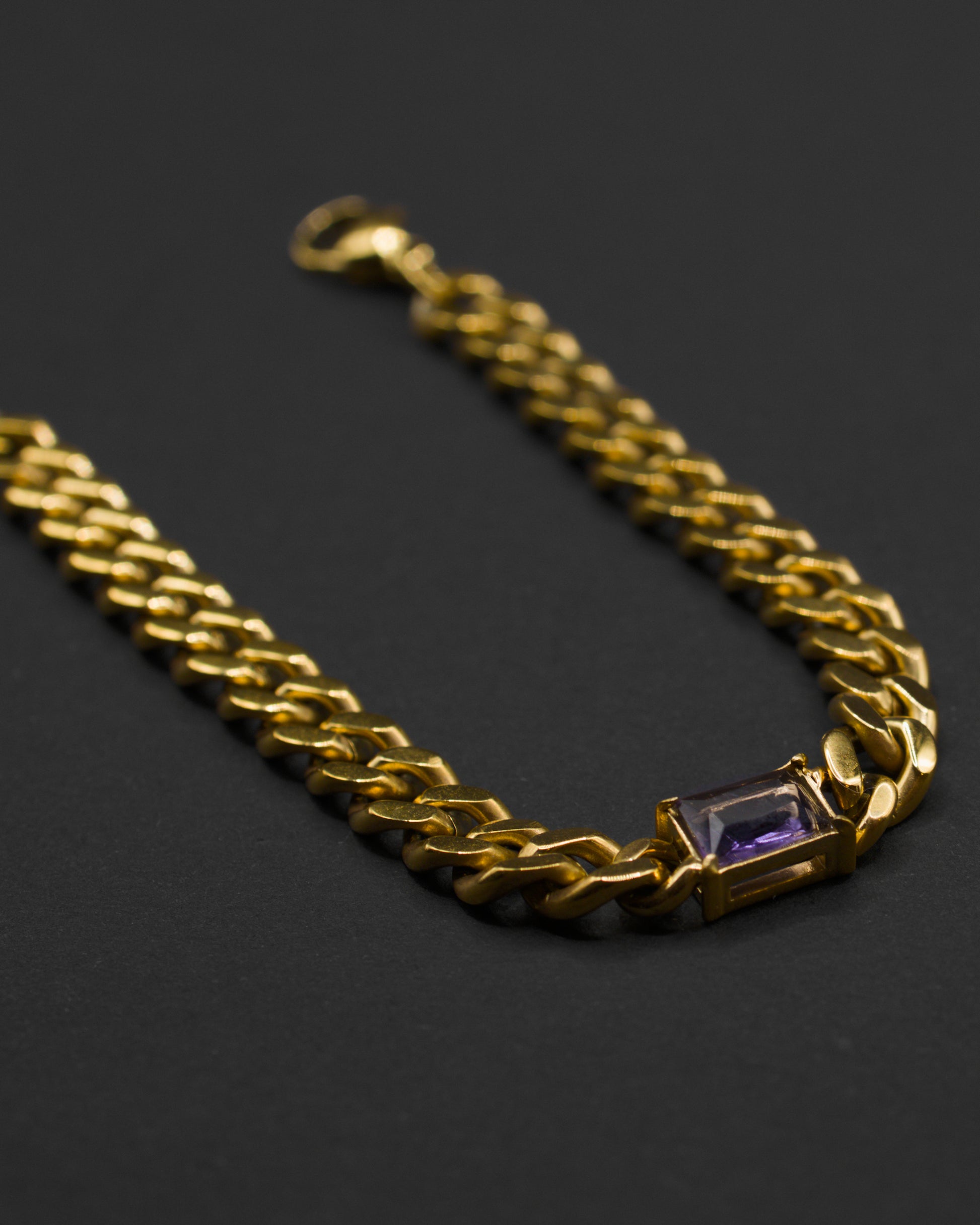Close-up of the gold Glowing Purple necklace in 18k gold plated 316L stainless steel, featuring a bold curb chain with a rectangular emerald-cut purple crystal pendant on a dark background.