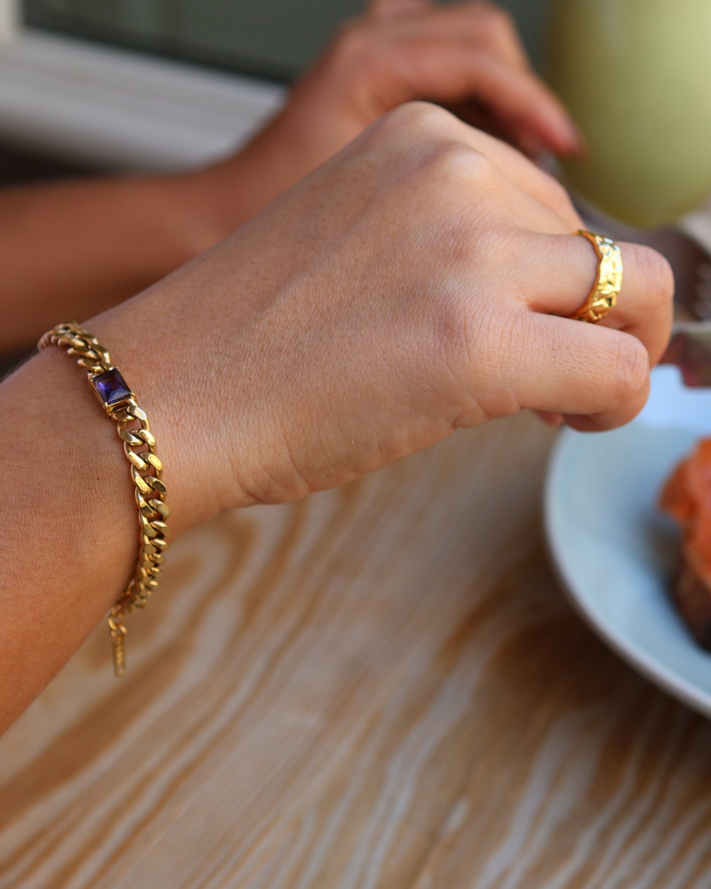 Close-up lifestyle shot of a woman's wrist wearing the gold Glowing Purple chain bracelet in 18k gold plated 316L stainless steel, featuring a rectangular purple crystal and styled with a textured gold ring.