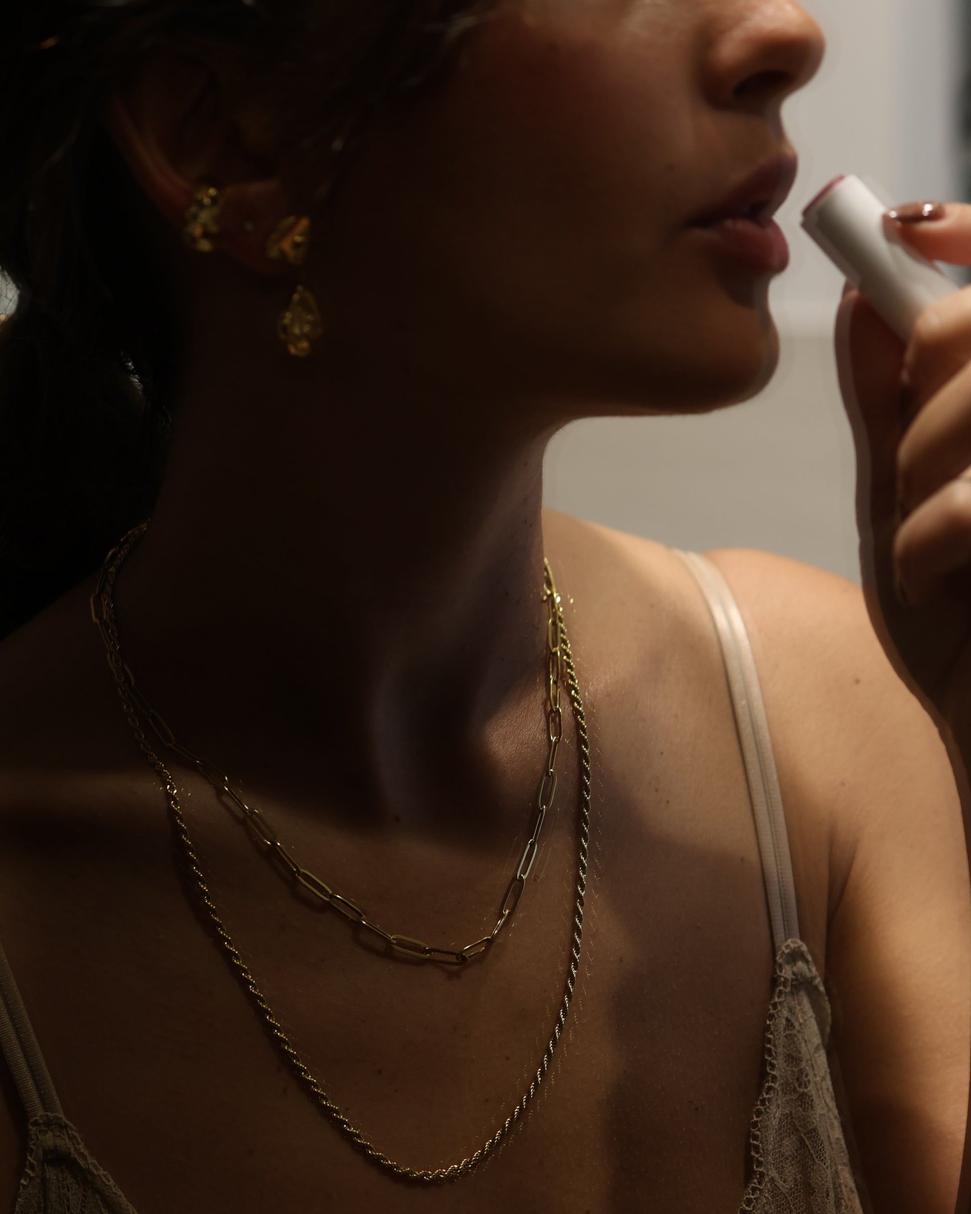 Lifestyle shot of a woman wearing a gold necklace stack featuring the Echo chain in 18k gold plated 316L stainless steel, paired with a thin rope chain and gold earrings in a warm, moody lighting.