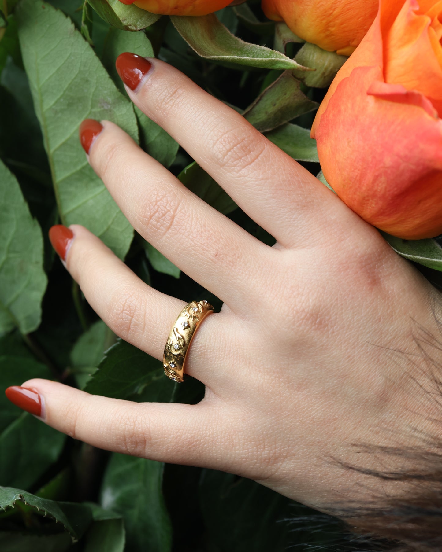 Close-up of a gold celestial band ring with sun symbols worn on a hand with red nails and green leaves.