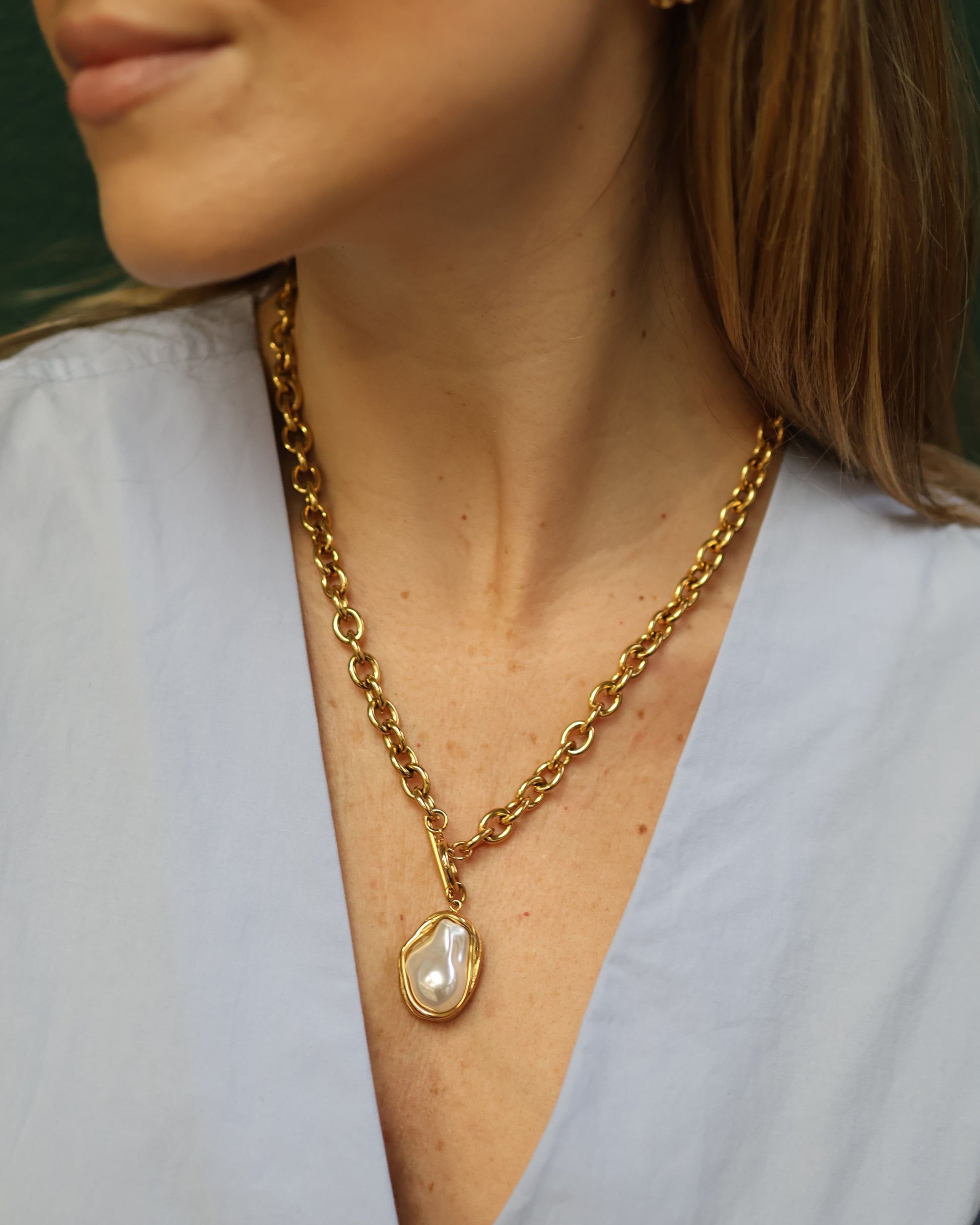 Close-up of a chunky gold chain necklace with round links and a large irregular baroque pearl pendant in a gold setting. The jewelry is styled over a light blue V-neck top, emphasizing the gold luster and organic pearl texture for a modern fashion look.
