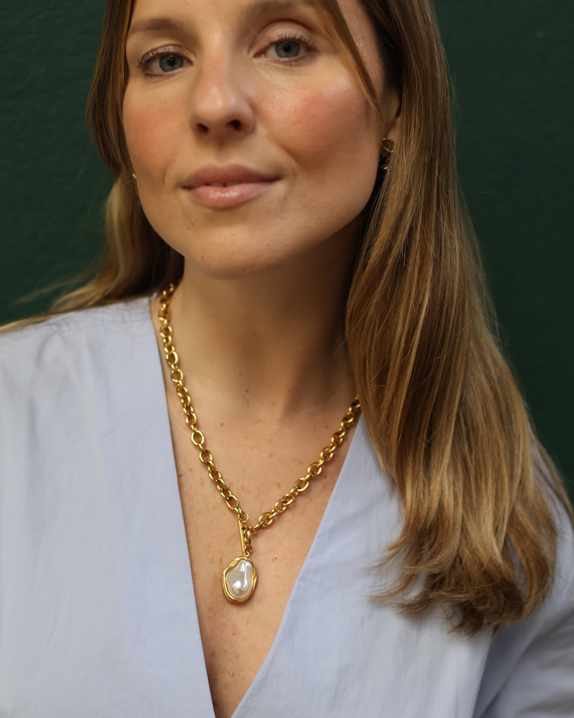 Smiling woman wearing a statement gold chain necklace with a large baroque pearl. Fashion portrait featuring modern gold jewelry, light blue blouse, and a dark green background for lifestyle SEO.