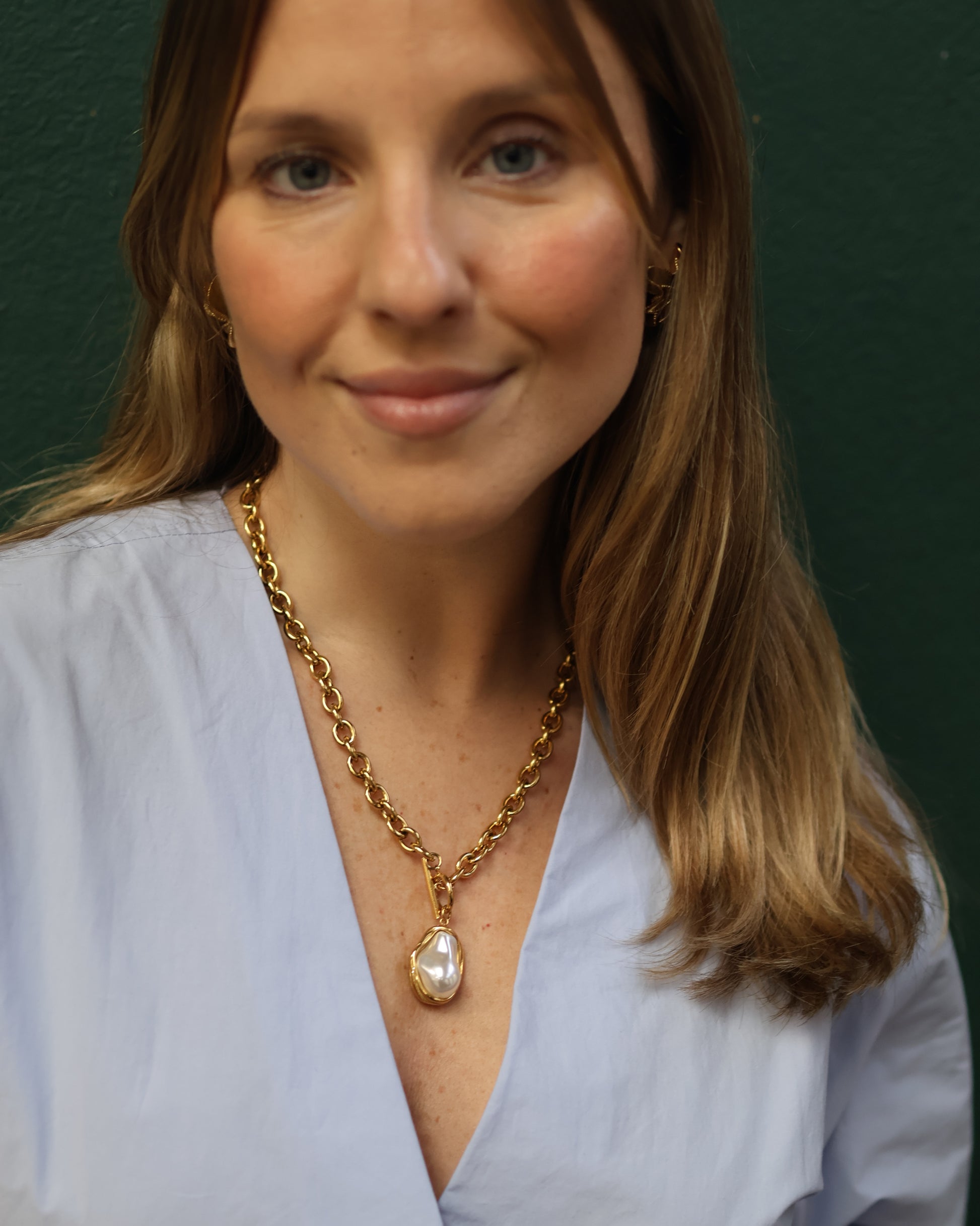 Close-up of a woman with wavy blonde hair smiling softly at the camera. She is wearing a light blue V-neck top and a statement gold chain necklace featuring a large baroque pearl pendant encased in gold. The background is a dark green textured wall.